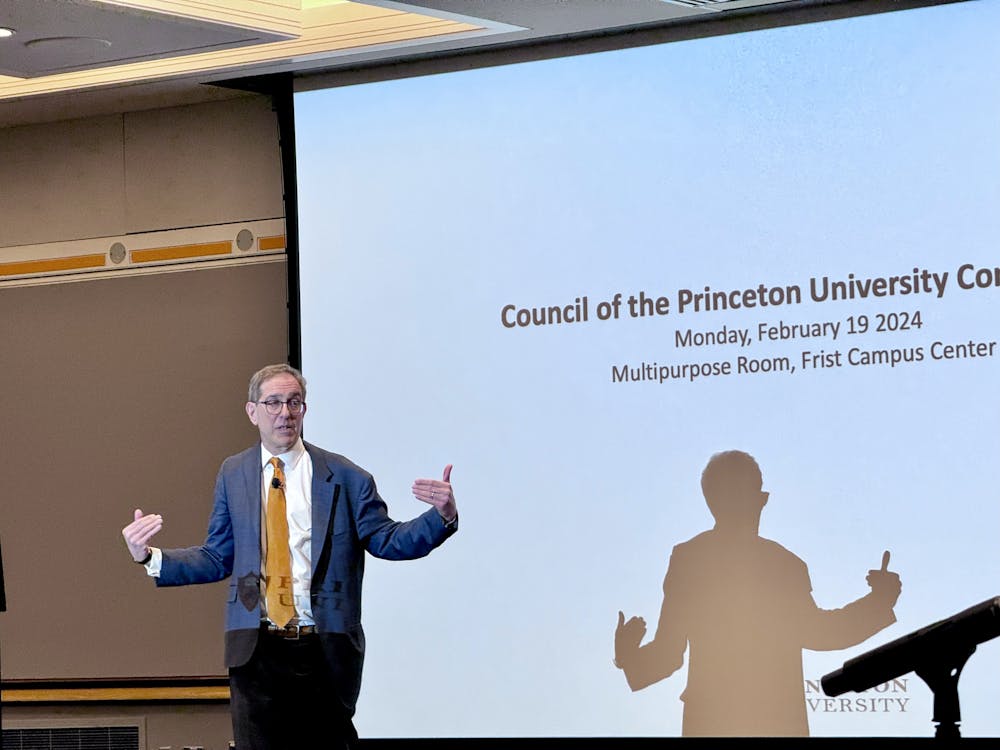 A man with glasses and an orange tie points at himself in front of a screen that says "Council of the Princeton University Community."
