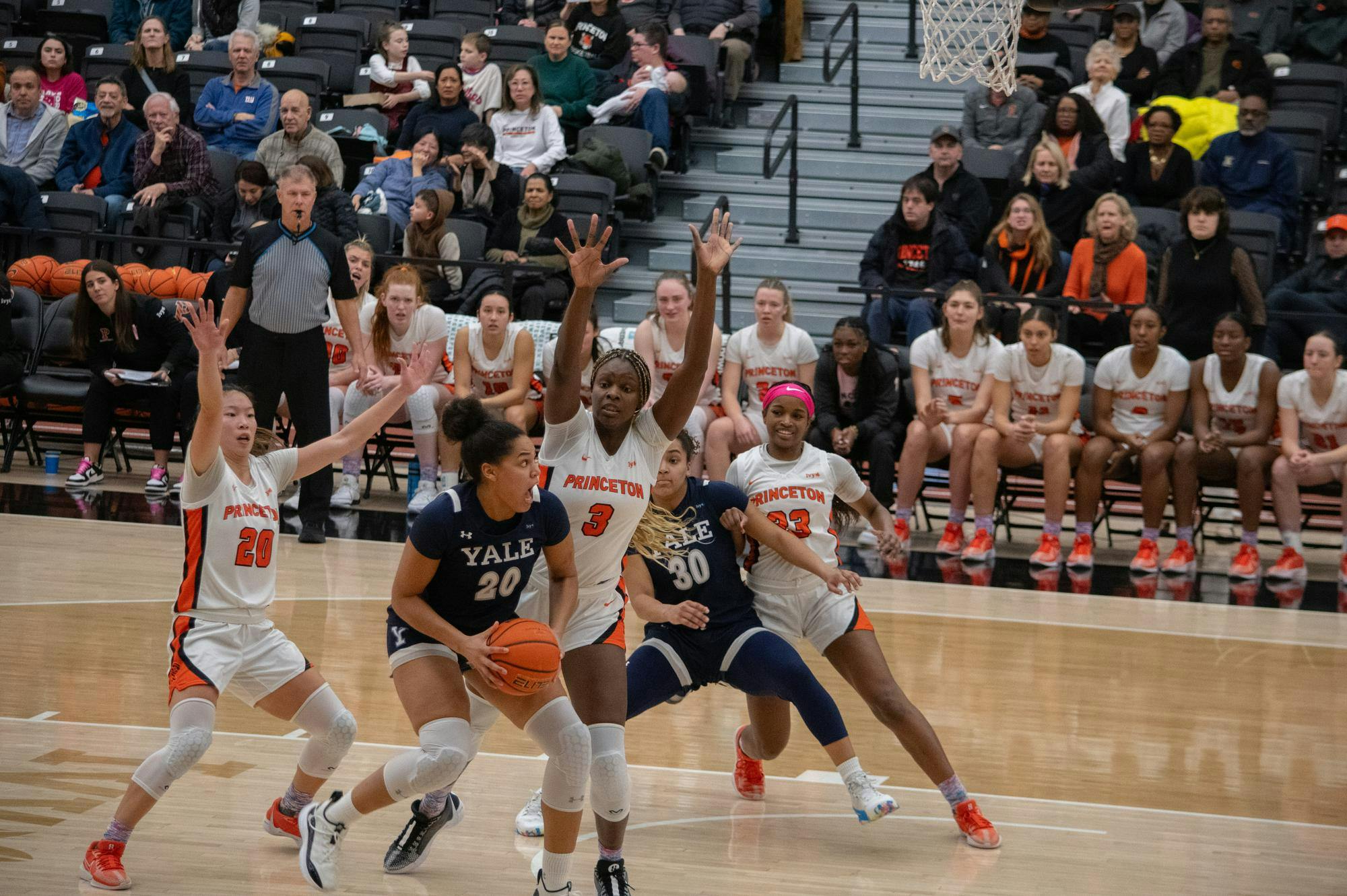 Two women, both wearing white and orange jerseys, block another woman wearing a blue and white jersey from shooting. They are standing on a tan court with a bench of players and the crowd in the background.