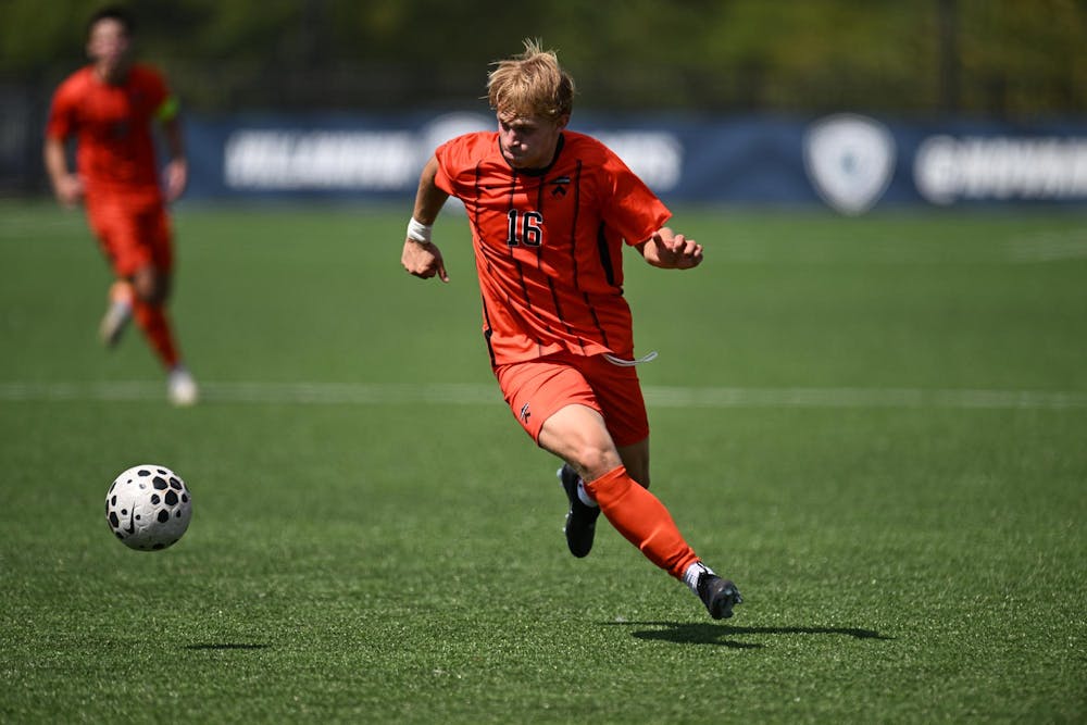 A man wearing orange shorts and an orange jersey with black vertical stripes on it as he runs towards a soccer ball on a turf field. 