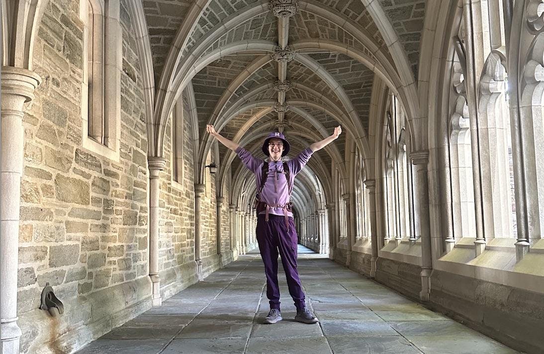 A young man wearing a purple hat, purple shirt, purple pants, and a purple backpack of varying shades stands with his arms in the air and a smile on his face in an arched stone outdoor walkway. 