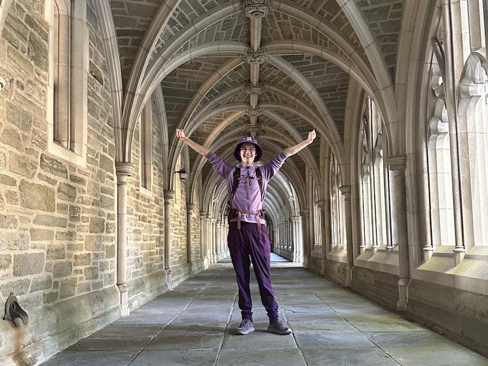 A young man wearing a purple hat, purple shirt, purple pants, and a purple backpack of varying shades stands with his arms in the air and a smile on his face in an arched stone outdoor walkway.