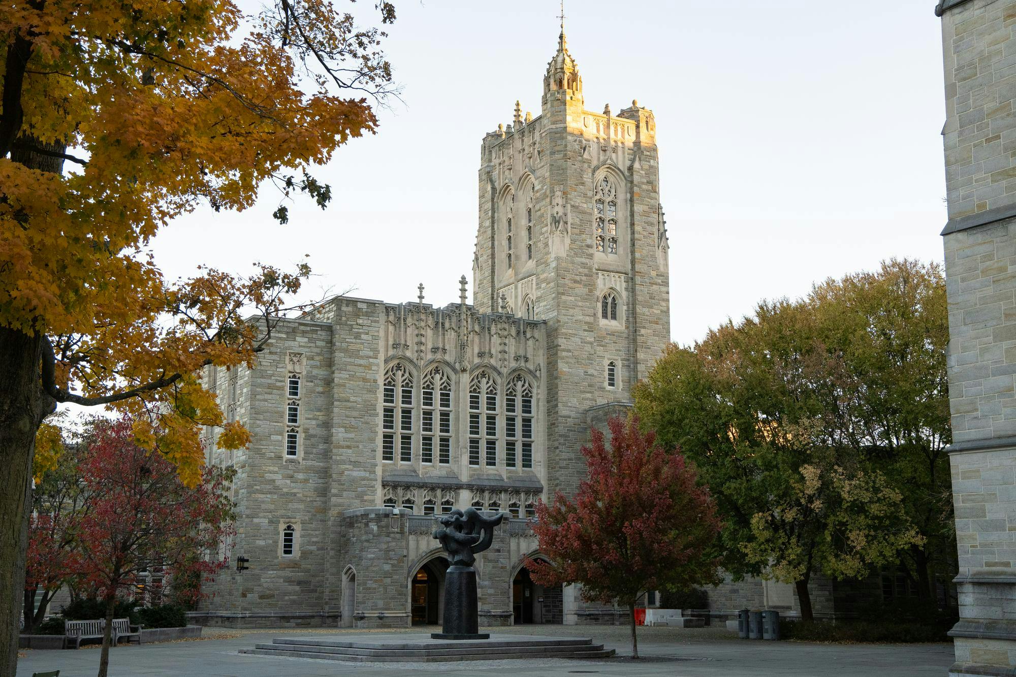 A stone building with a tall tower and intricate windows stands behind a black statue and an array of autumn trees. 