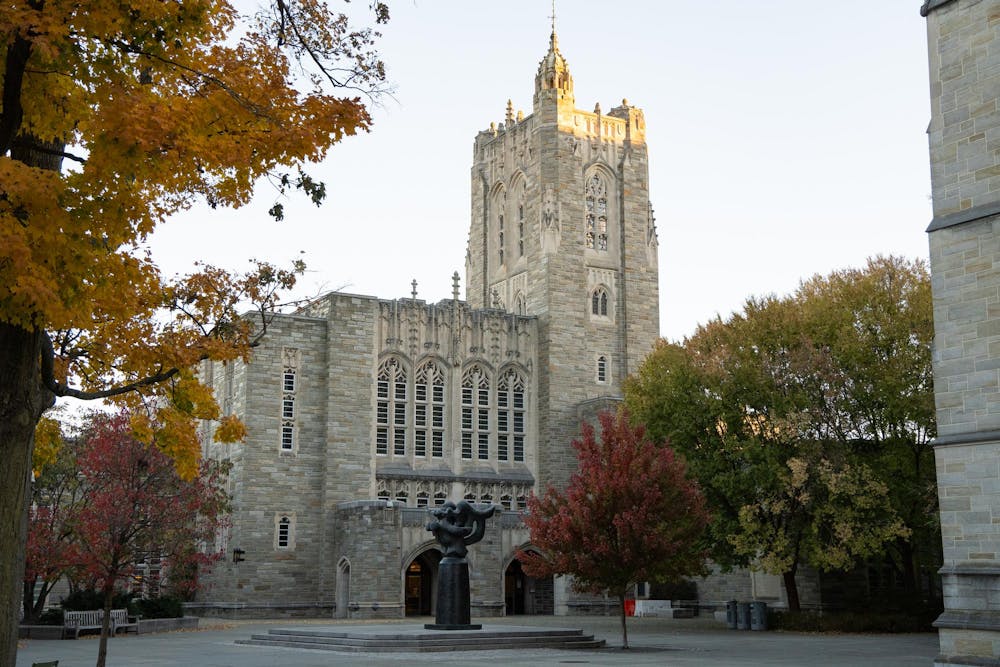 A stone building with a tall tower and intricate windows stands behind a black statue and an array of autumn trees. 