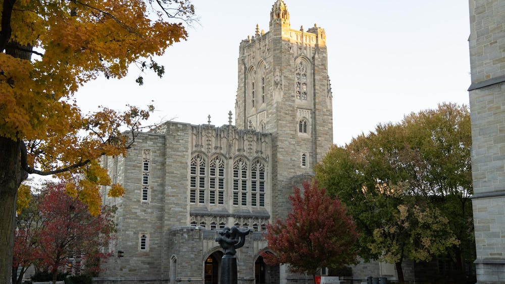 A stone building with a tall tower and intricate windows stands behind a black statue and an array of autumn trees.