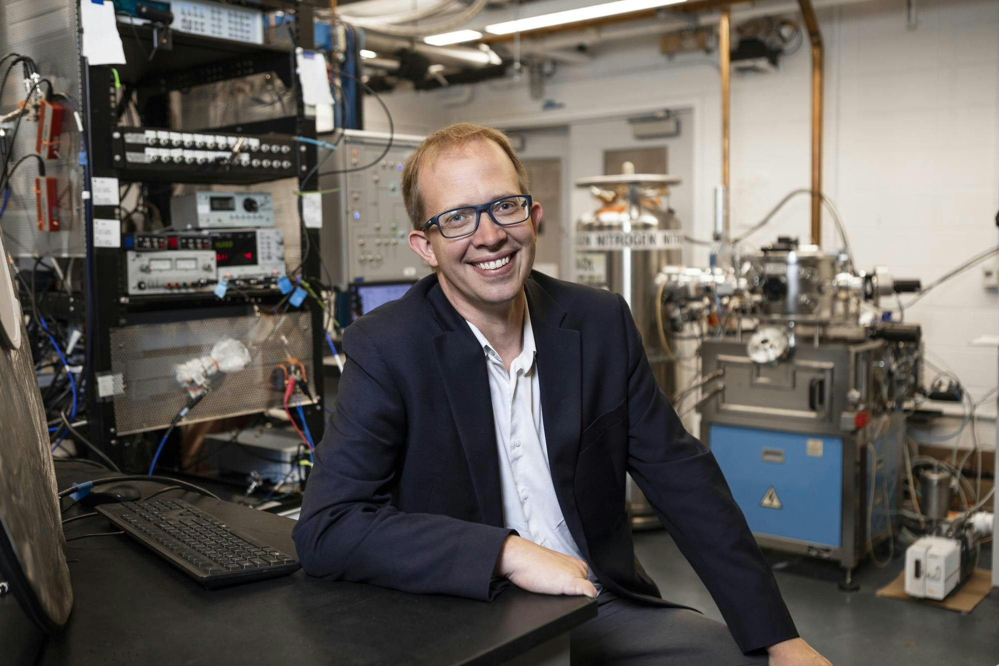 Man wearing glasses and a navy suit sits in a science laboratory.