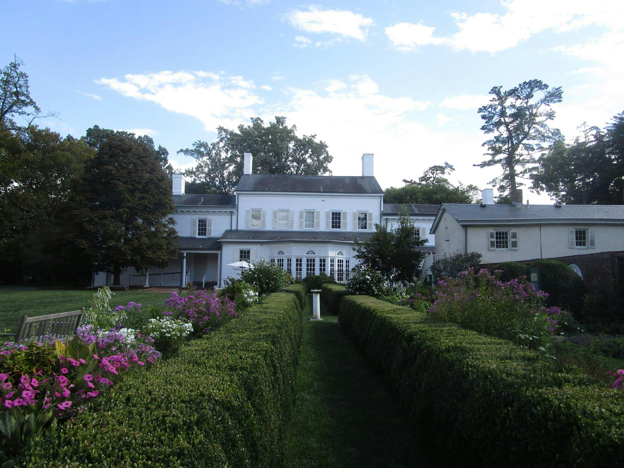 A white mansion with a bay window sits behind a hedge-rowed, manicured green garden with white and purple flowers. 