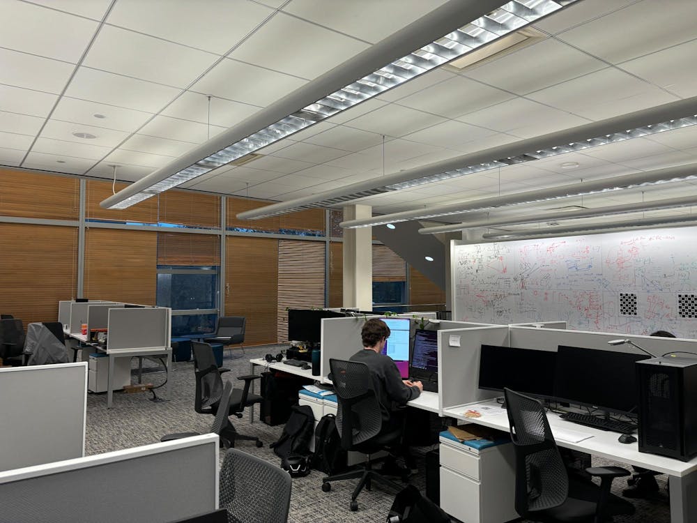 Gray cubicles with computers are lined up in rows in a basement room. 