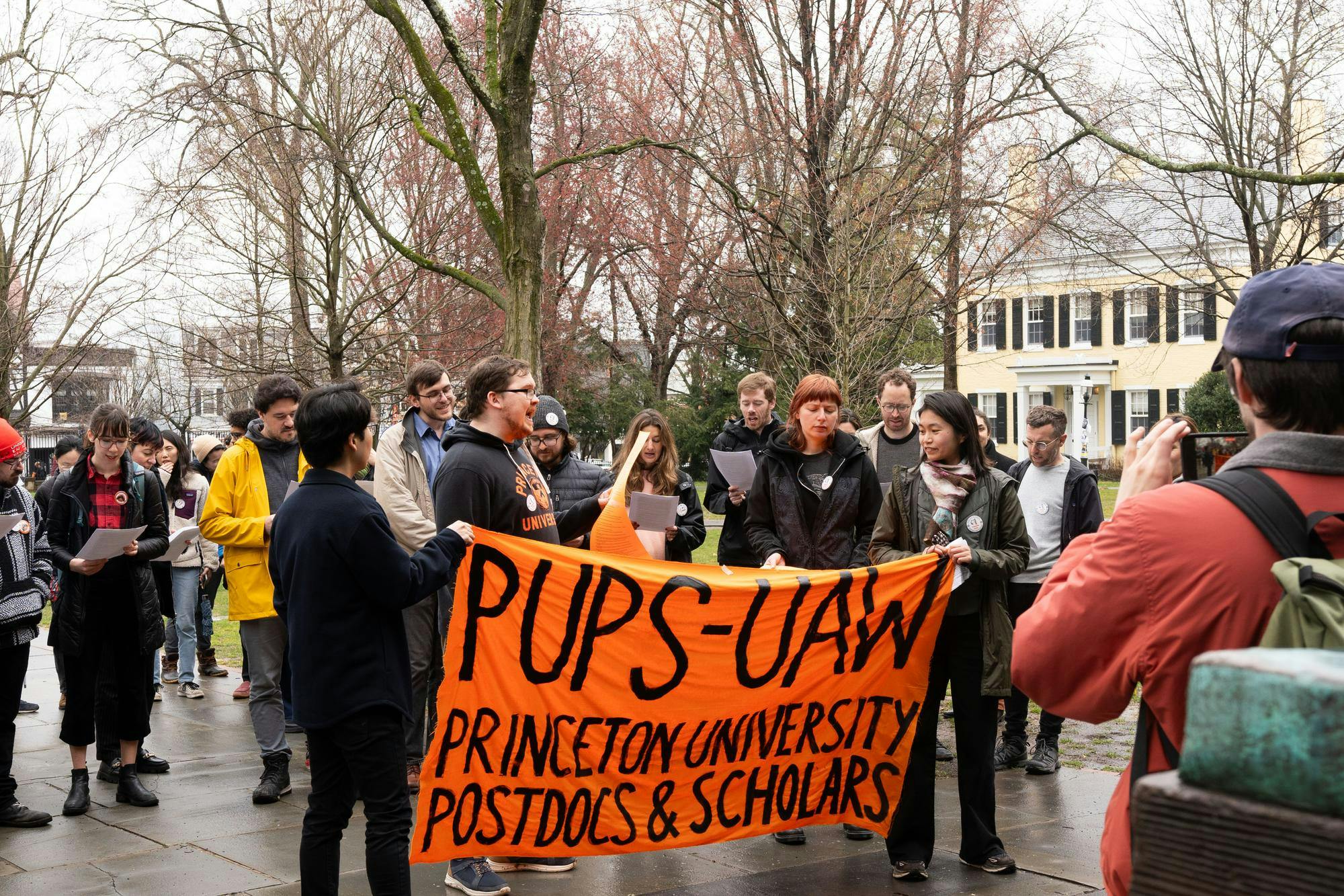 A crowd of several dozen people hold a large orange banner that reads “PUPS-UAW PRINCETON UNIVERSITY POSTDOCS AND SCHOLARS”
