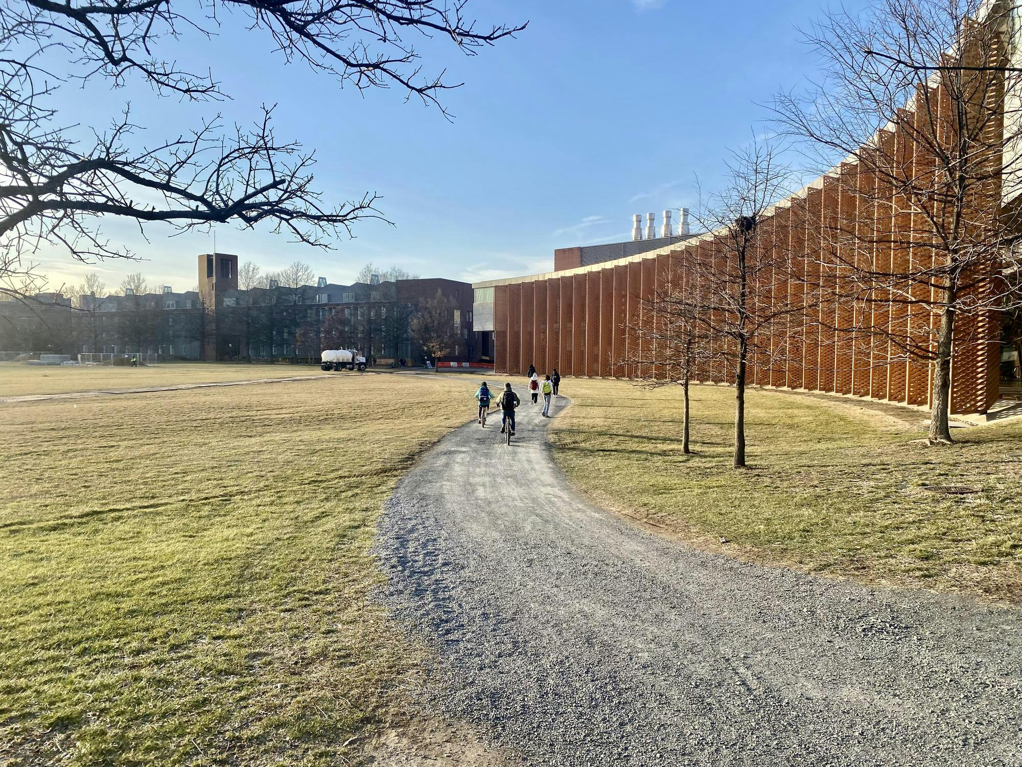 On the right, a red-hued building with metal flaps for shade, behind a gravel path with several bikers. Grass on the left.