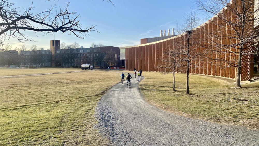 On the right, a red-hued building with metal flaps for shade, behind a gravel path with several bikers. Grass on the left.