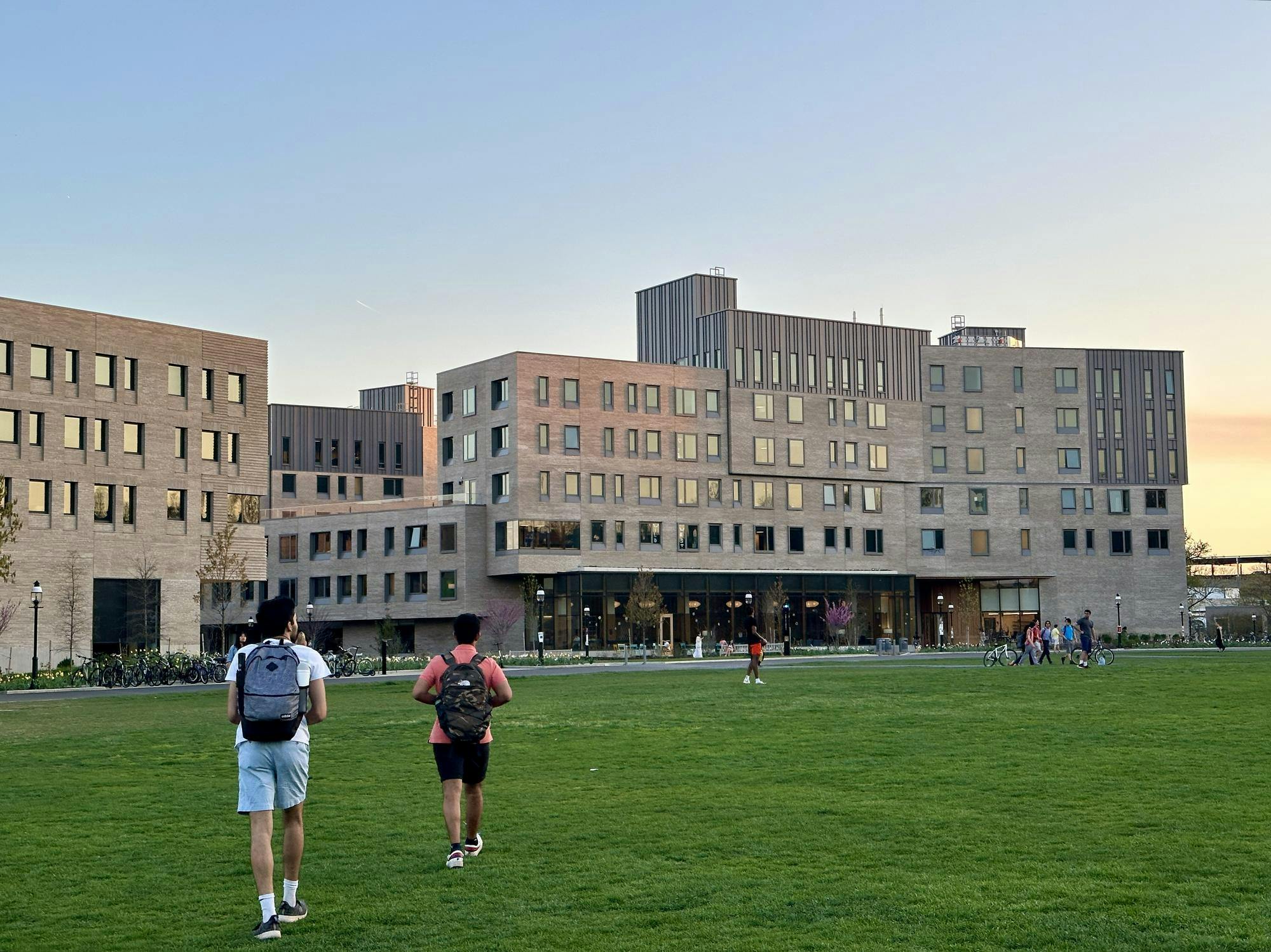 Two people with backpacks walk across a green field towards a series of blocky gray buildings