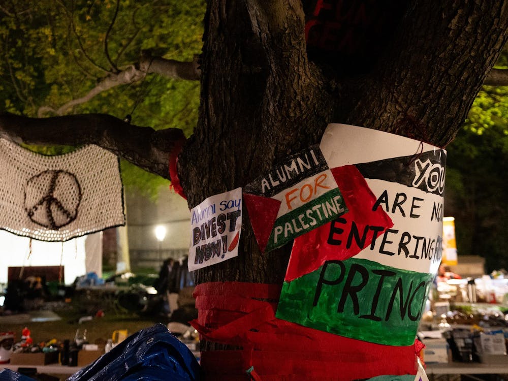 Tree wrapped in various posters in support of Palestine, and red streamer is wrapped around the space below. A crocheted peace symbol is hanging from a branch.