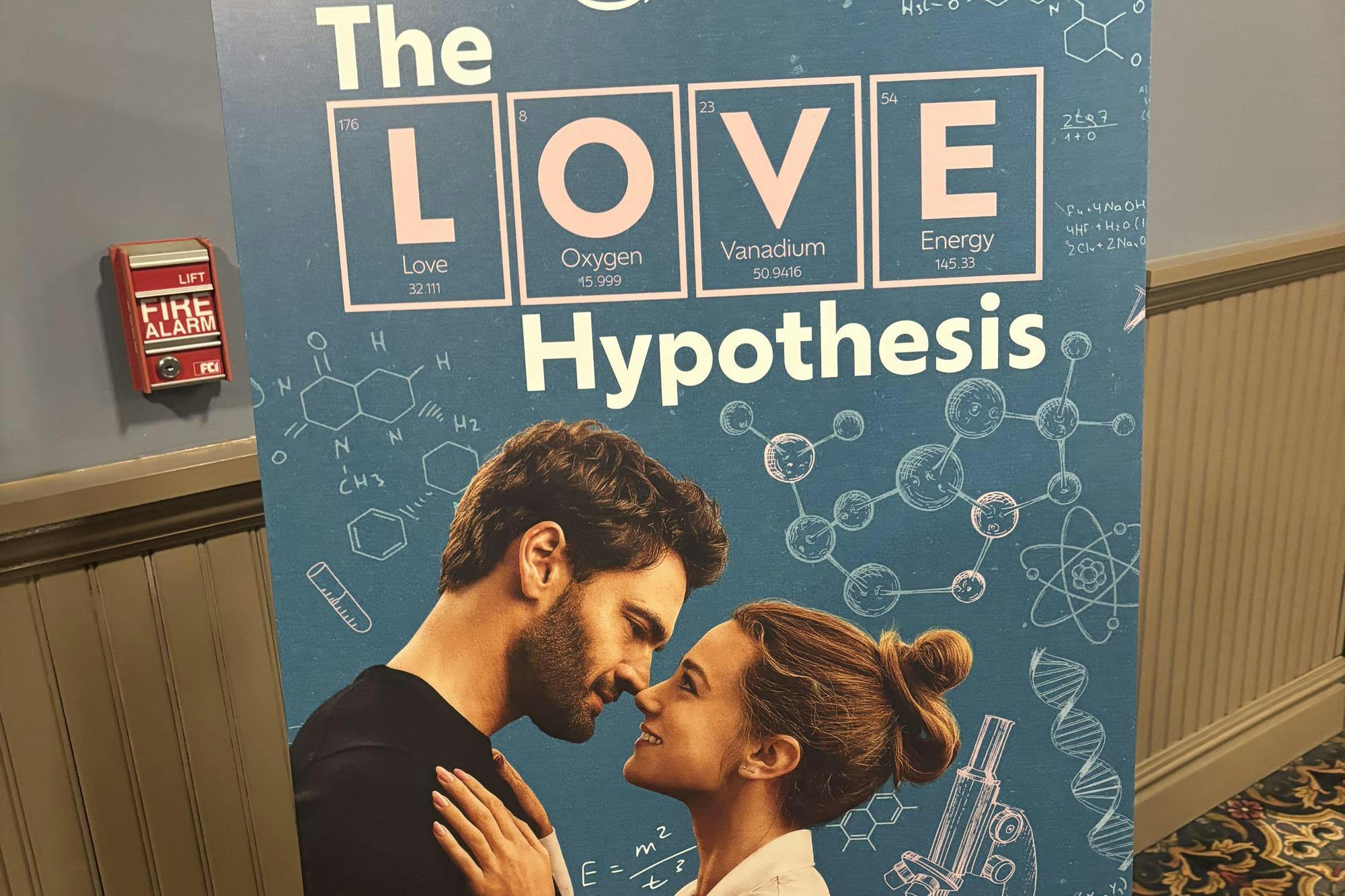 A movie poster with a blue background is seen against a gray wall, titled "The Love Hypothesis," with a woman and a man holding each other and looking at one another. 