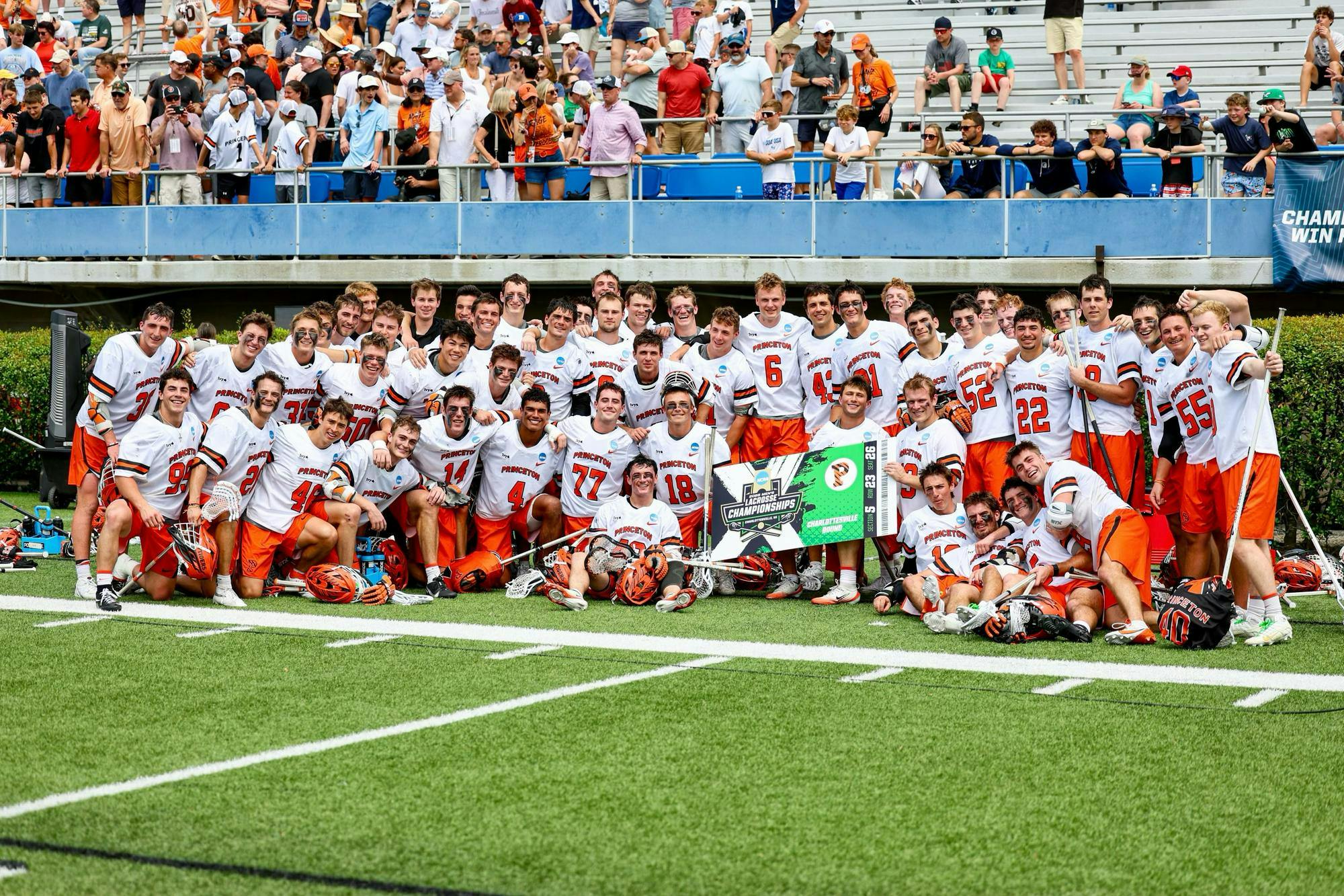 A group of Princeton lacrosse players in orange shorts and white jerseys posing for a photo with a banner that reads “Charlottesville Bound.” 