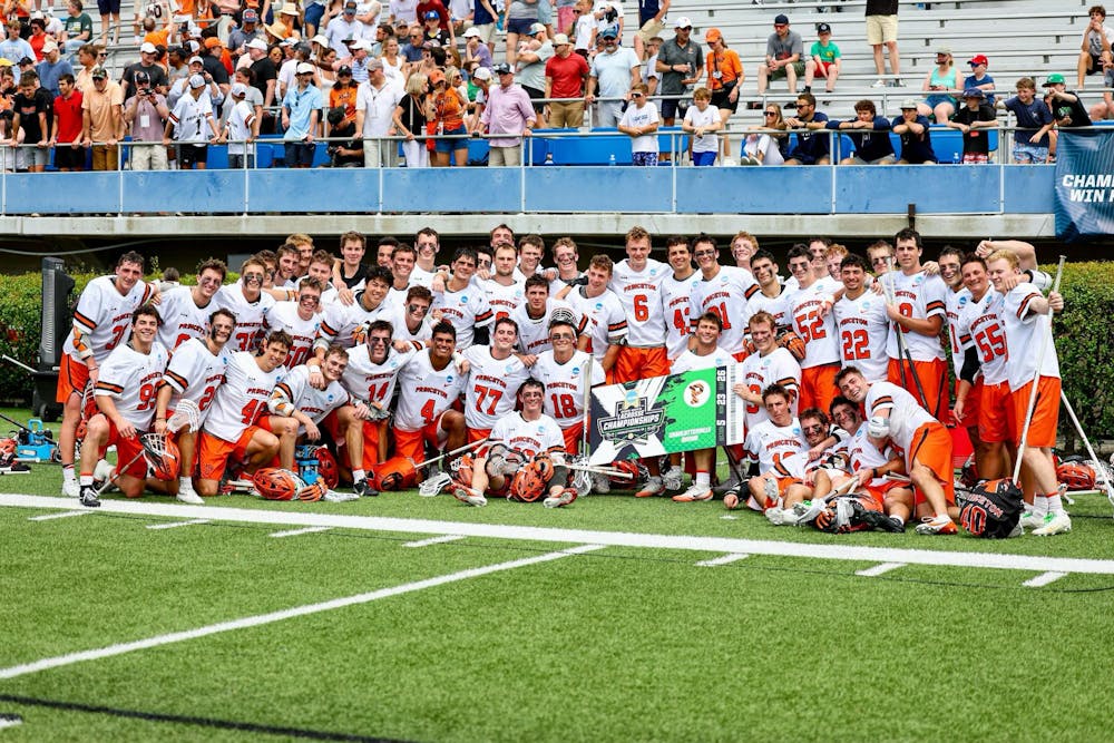 A group of Princeton lacrosse players in orange shorts and white jerseys posing for a photo with a banner that reads “Charlottesville Bound.” 
