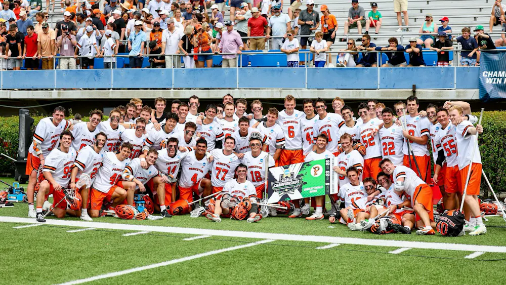 A group of Princeton lacrosse players in orange shorts and white jerseys posing for a photo with a banner that reads “Charlottesville Bound.”