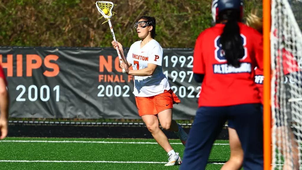 Girl in white shirt and orange skirt cradles a lacrosse ball in her stick.