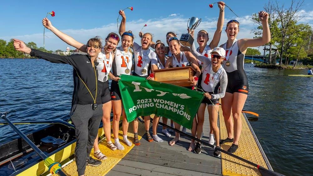 Ten women stand on a boat dock with roses in hand all holding large green flag reading 2023 Women’s Rowing Champions.