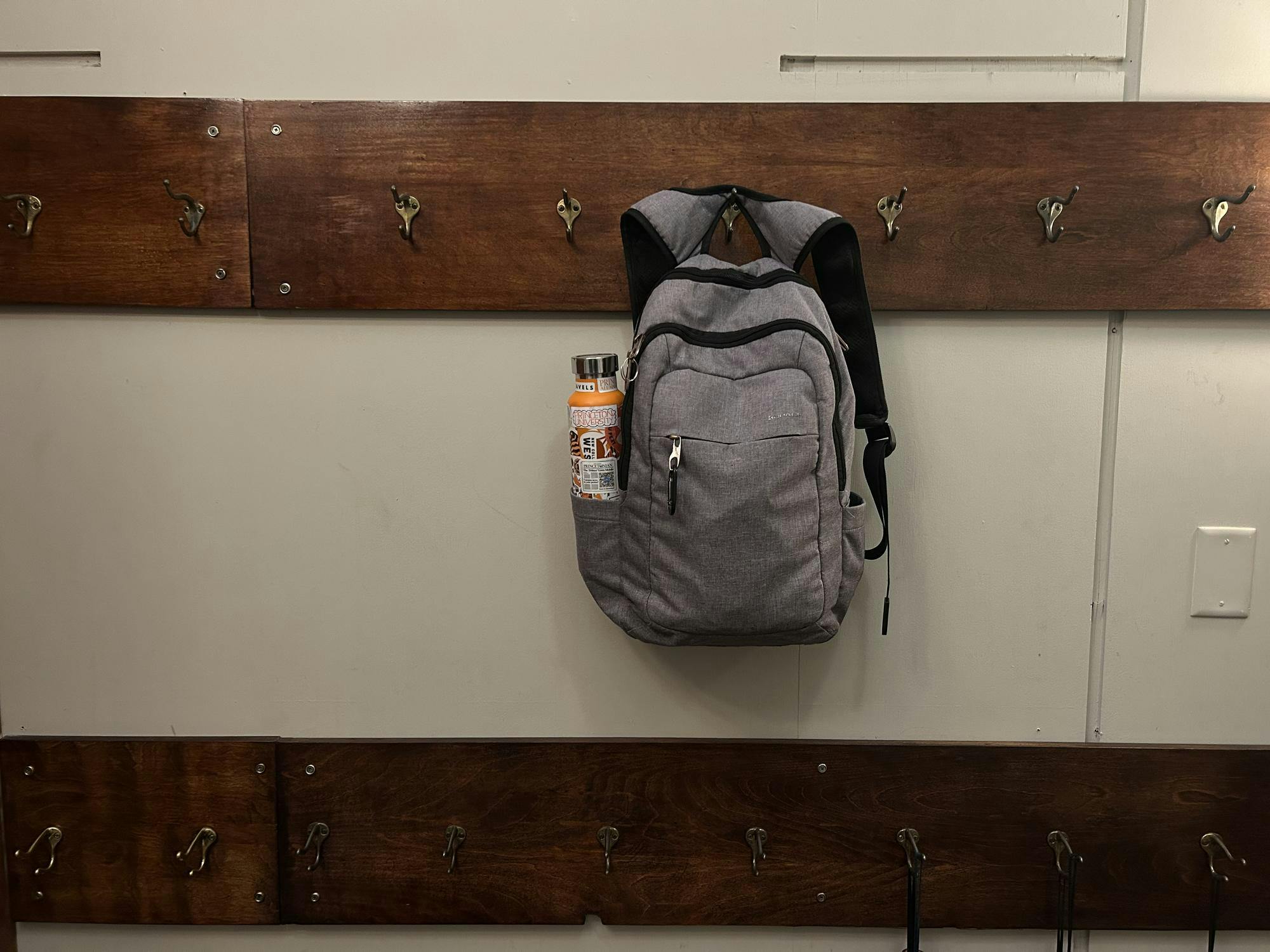 A gray backpack with an orange water bottle hangs from a metal hook. The gray metal hook is one of several gray metal hooks that are in a straight line and bolted to a long piece of brown wood. Another brown wooden panel with metal hooks is two feet underneath it.