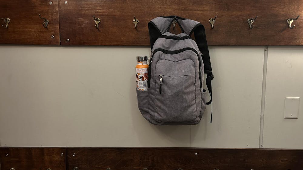 A gray backpack with an orange water bottle hangs from a metal hook. The gray metal hook is one of several gray metal hooks that are in a straight line and bolted to a long piece of brown wood. Another brown wooden panel with metal hooks is two feet underneath it.