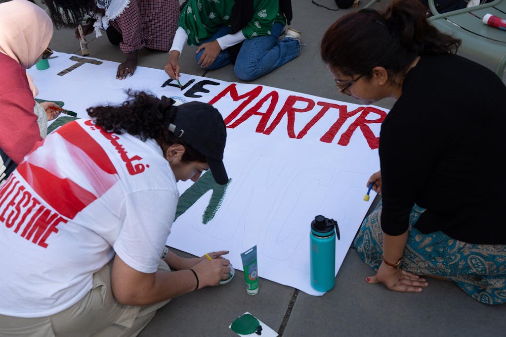 A group of individuals is seated on the ground, painting a large protest banner. The banner features the word "MARTYR" in big red letters. The other letters, still being painted in black and green, are part of a larger message. One participant, wearing a white t-shirt with a red graphic that reads "Palestine" in English and Arabic, focuses on painting a section of the banner in green. Another person,, works on a different part of the banner. Paint supplies and water bottles are scattered around the group as work.