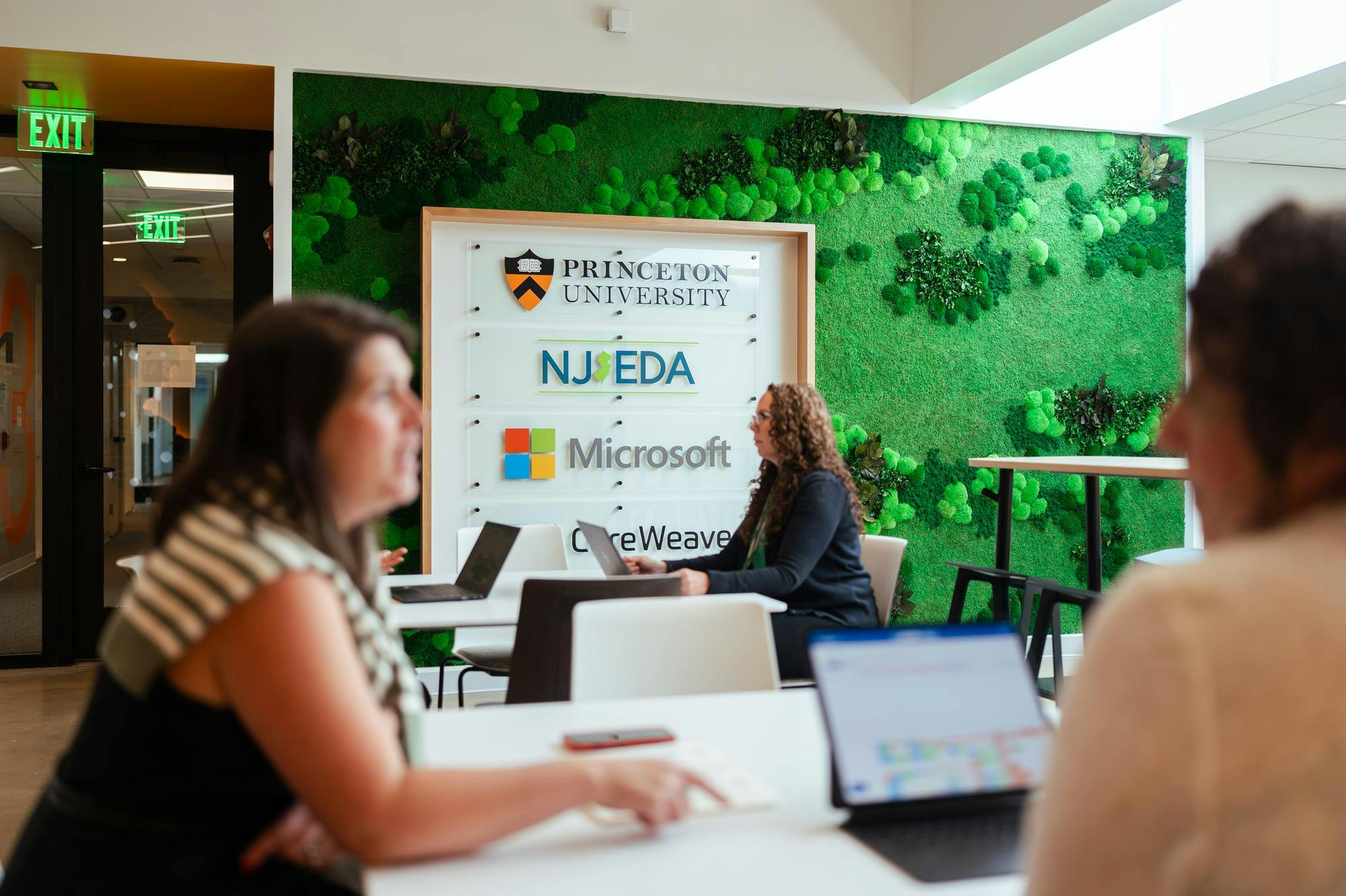 Focused on a woman working on her computer, sitting in front of the AI Hub Founding Partners sign. 