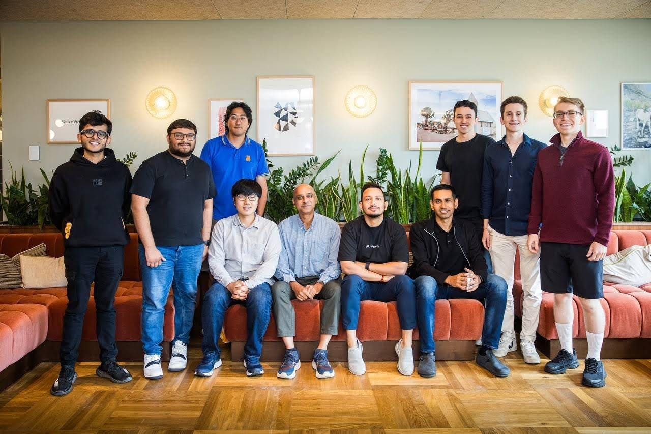A group of ten people with four sitting on a red couch and three standing to either side, all in front of houseplants and paintings.