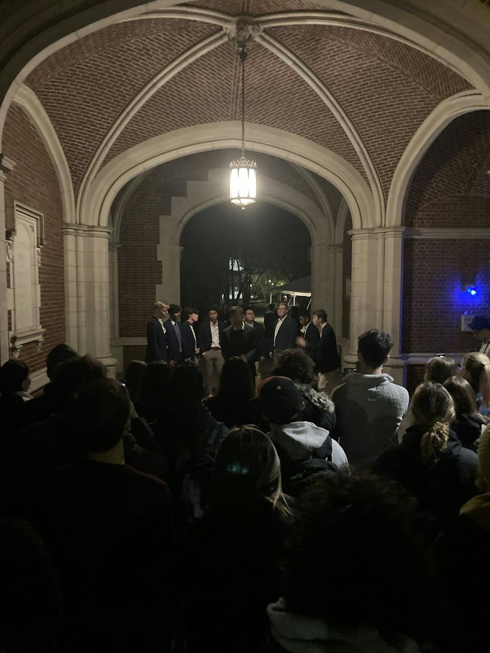 In an arch, at night, a group of formally-dressed men stand in a semi-circle.