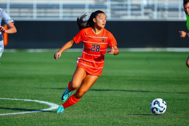 A woman wearing an orange jersey on a grass field during a soccer match. 
