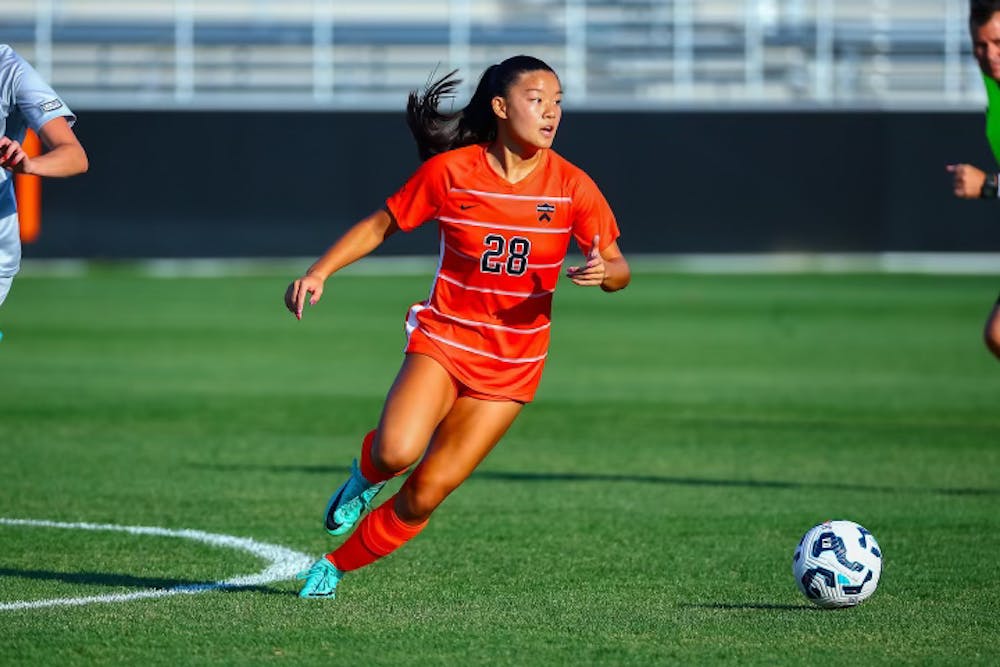 A woman wearing an orange jersey on a grass field during a soccer match. 