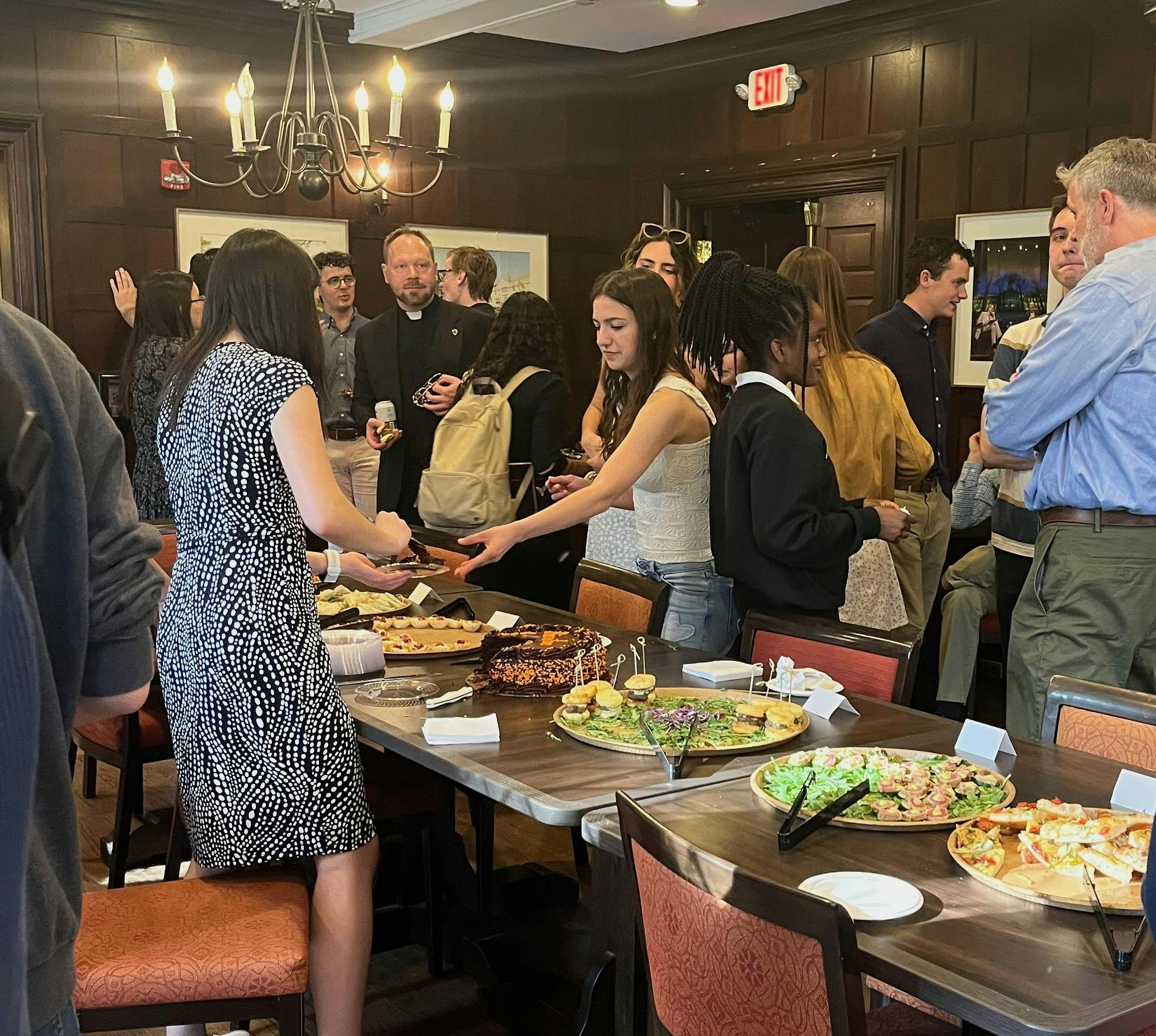 A group of people is standing around a table. Some are chatting, and others are eating food. 