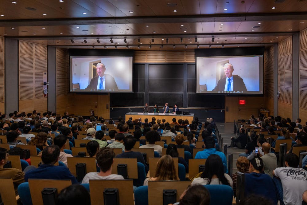 A large lecture hall is filled with an audience seated in rows. At the front of the room, a panel of four speakers is seated behind a desk. Two large screens above the panel display a virtual speaker, an older man participating in the event via video conference. There are wood-paneled walls and high ceilings and a chalkboard is visible. 