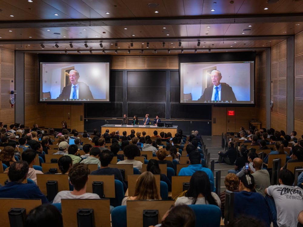 A large lecture hall is filled with an audience seated in rows. At the front of the room, a panel of four speakers is seated behind a desk. Two large screens above the panel display a virtual speaker, an older man participating in the event via video conference. There are wood-paneled walls and high ceilings and a chalkboard is visible.