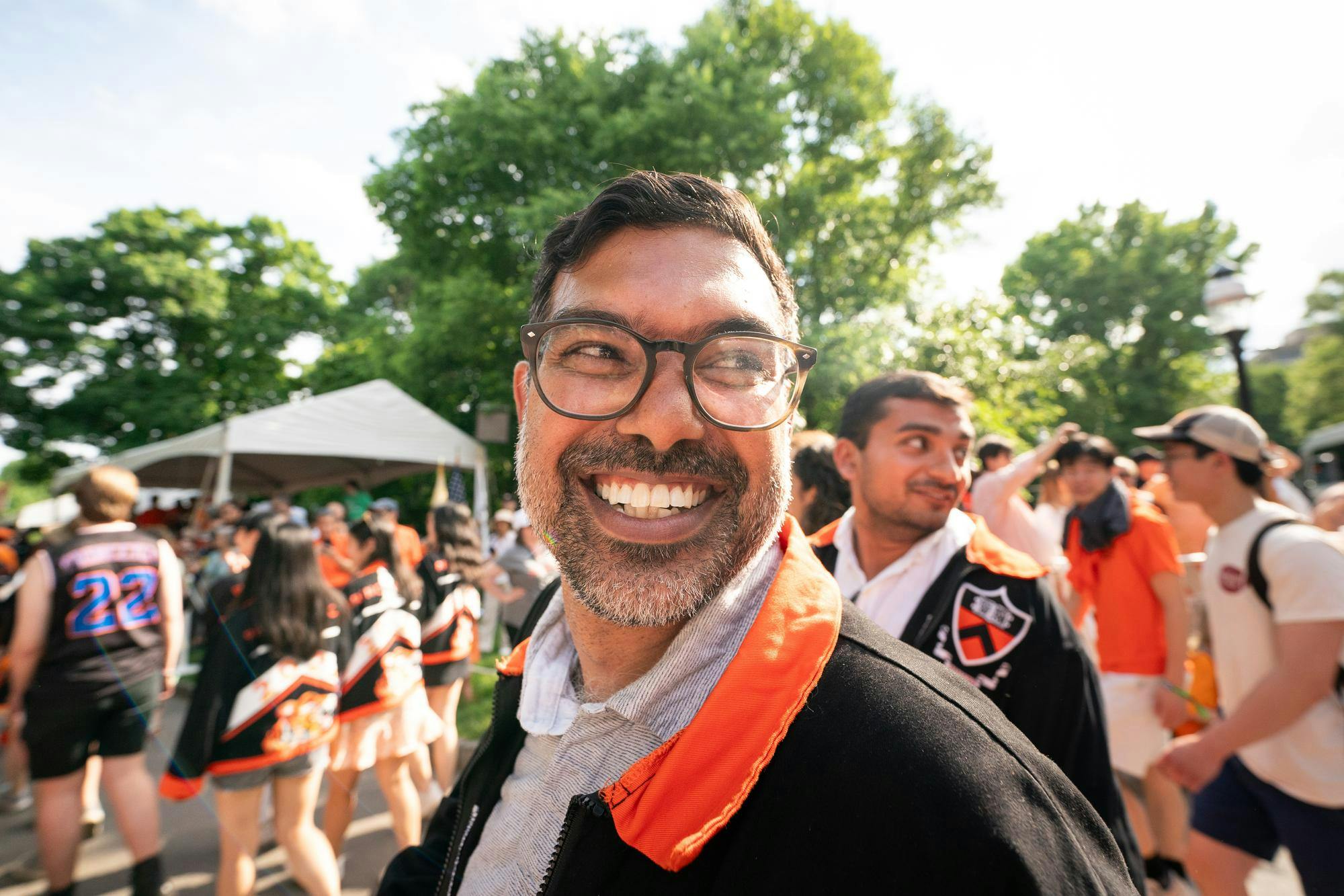 An Indian man with glasses smiles widely at the camera while looking right, with a large crowd of people behind him.