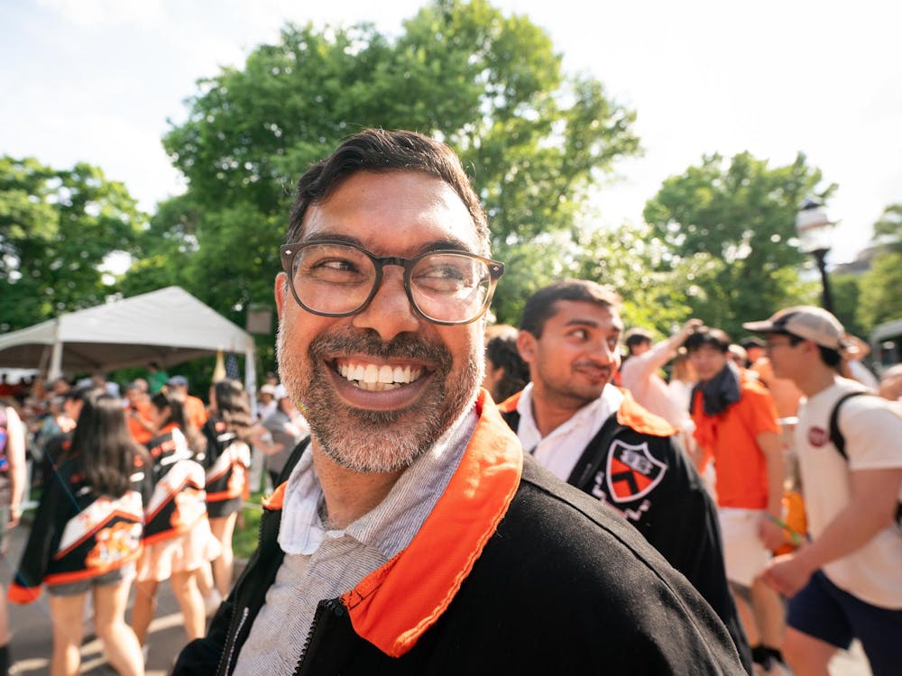 An Indian man with glasses smiles widely at the camera while looking right, with a large crowd of people behind him.