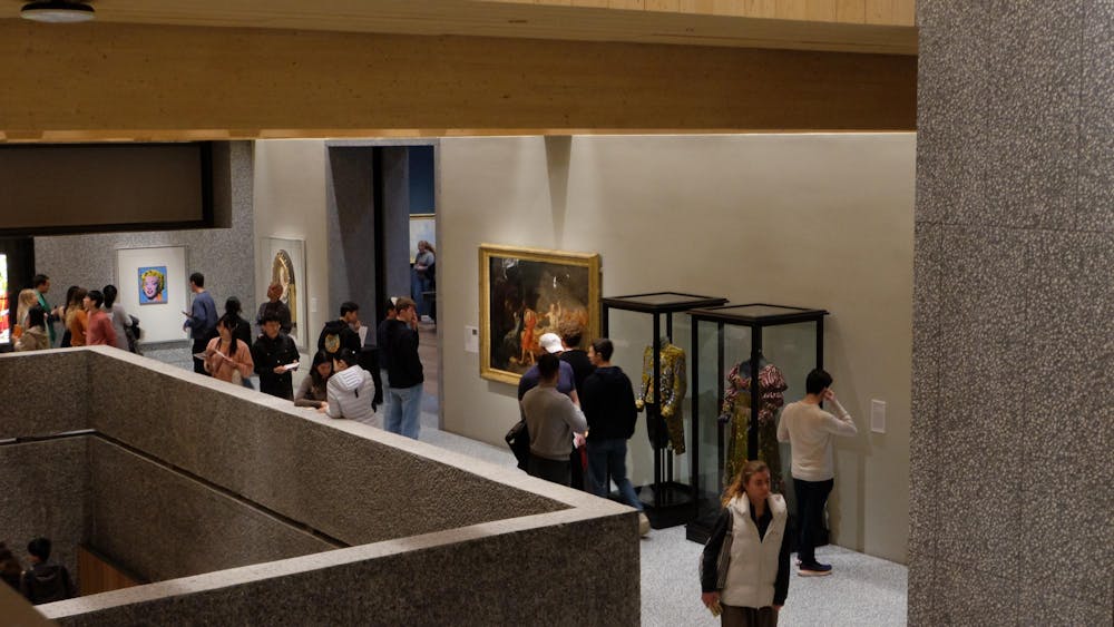 An elevated view of a bustling museum passageway. To the right, two costumes and a painting are against the wall. To the left, two paintings hang on the wall.
