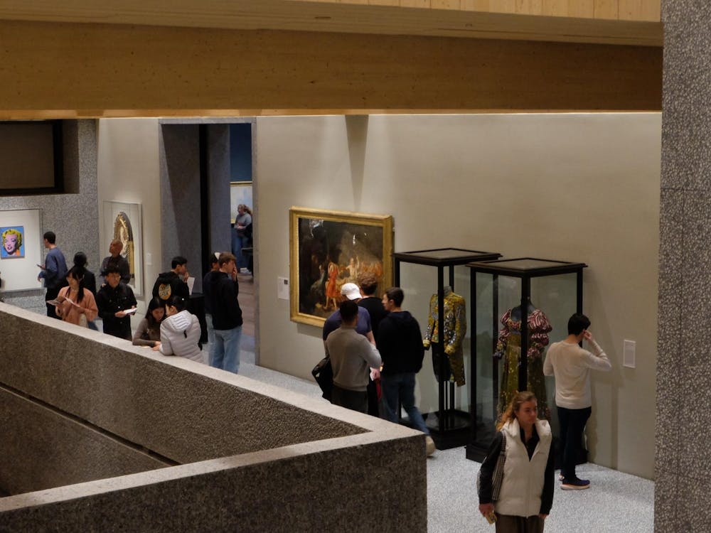 An elevated view of a bustling museum passageway. To the right, two costumes and a painting are against the wall. To the left, two paintings hang on the wall.