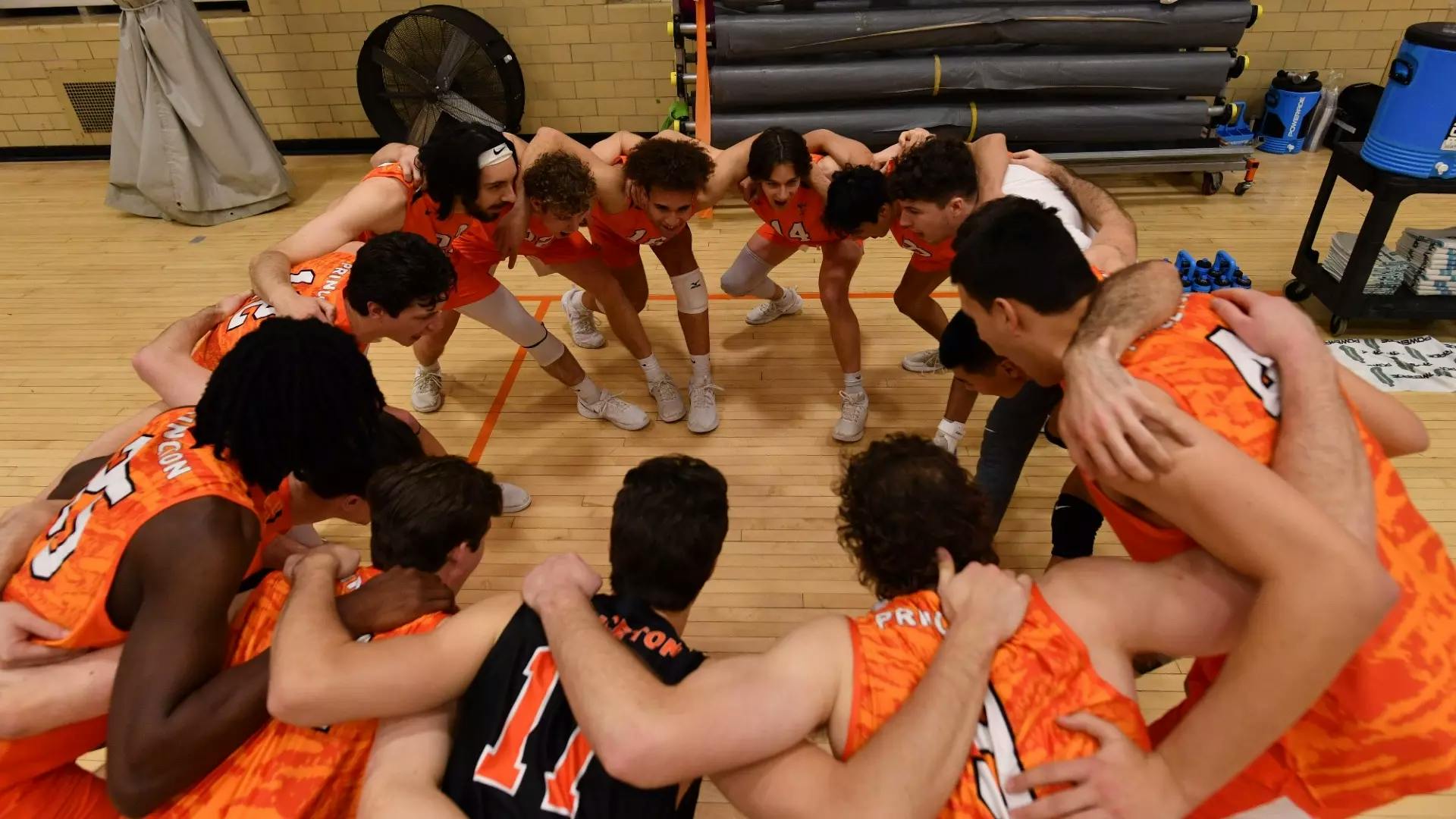 Players in orange and black gather in pregame huddle, with team captain the only player in black uniform. 