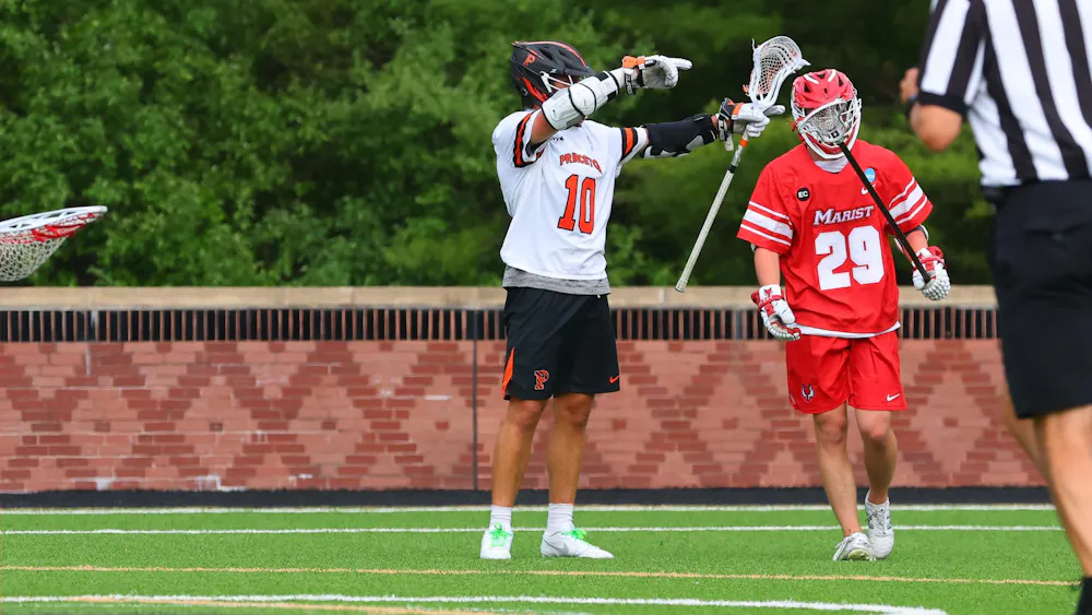 A Princeton lacrosse player wearing black shorts and a white No.10 jersey celebrates on a turf field with a Marist player in red shorts and a red jersey standing beside him.