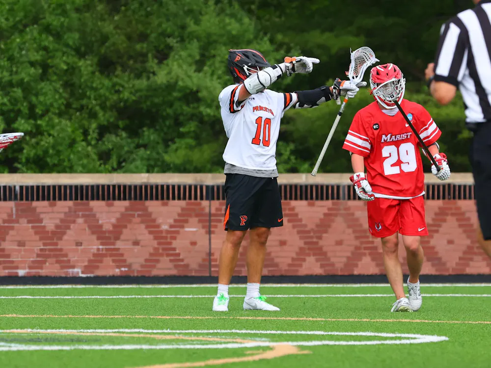 A Princeton lacrosse player wearing black shorts and a white No.10 jersey celebrates on a turf field with a Marist player in red shorts and a red jersey standing beside him.