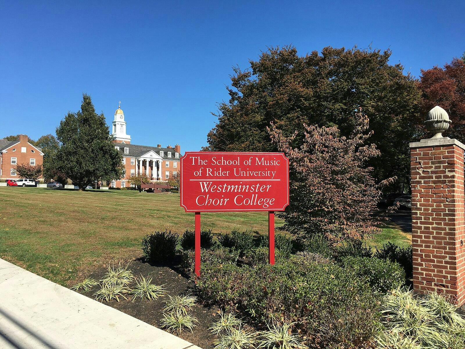 A red sign stands at the forefront. It reads: “The School of Music of Rider University; Westminster Choir College.” In the background are large trees and a colonial style building. 