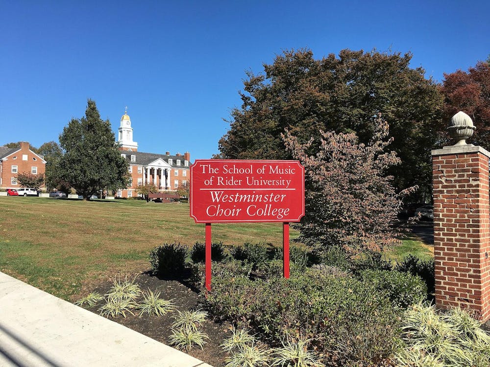 A red sign stands at the forefront. It reads: "The School of Music of Rider University; Westminster Choir College." In the background are large trees and a colonial style building. 