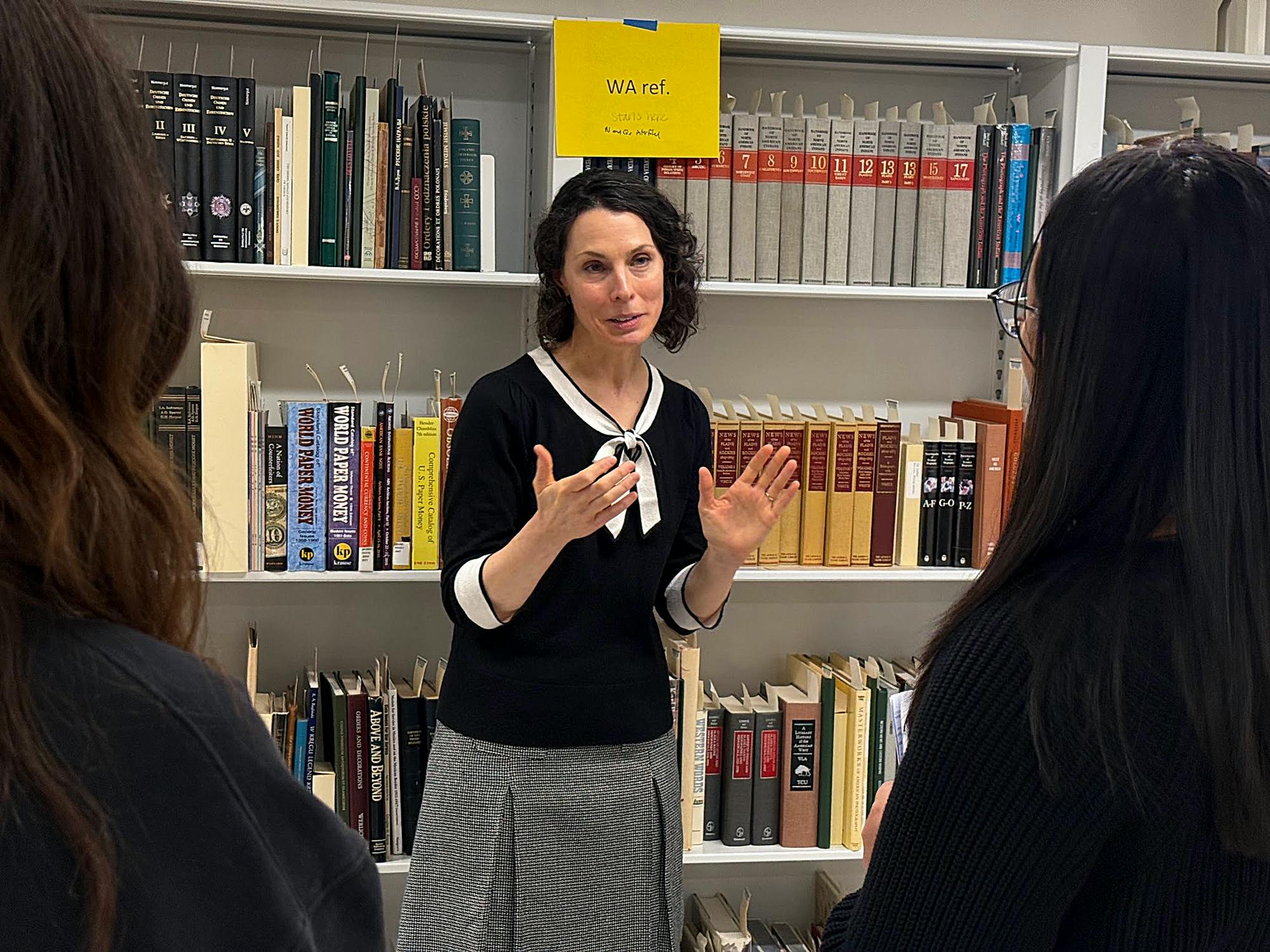 A woman with short dark hair wearing a black shirt with white trim stands before shelves of books, speaking with two women who stand facing her.