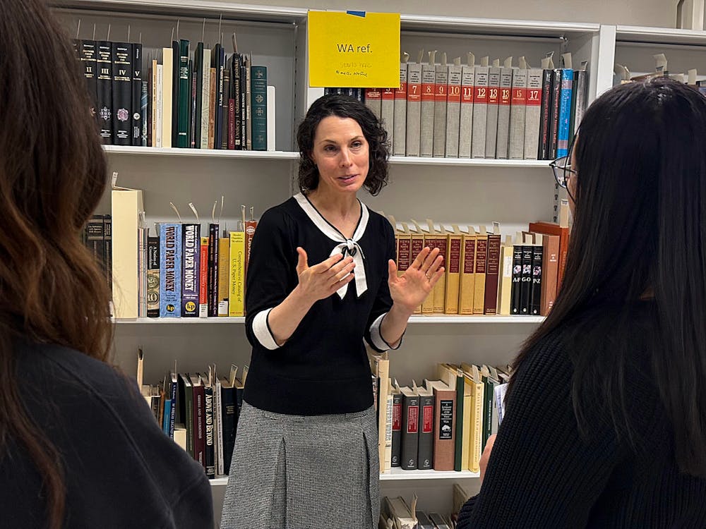 A woman with short dark hair wearing a black shirt with white trim stands before shelves of books, speaking with two women who stand facing her.
