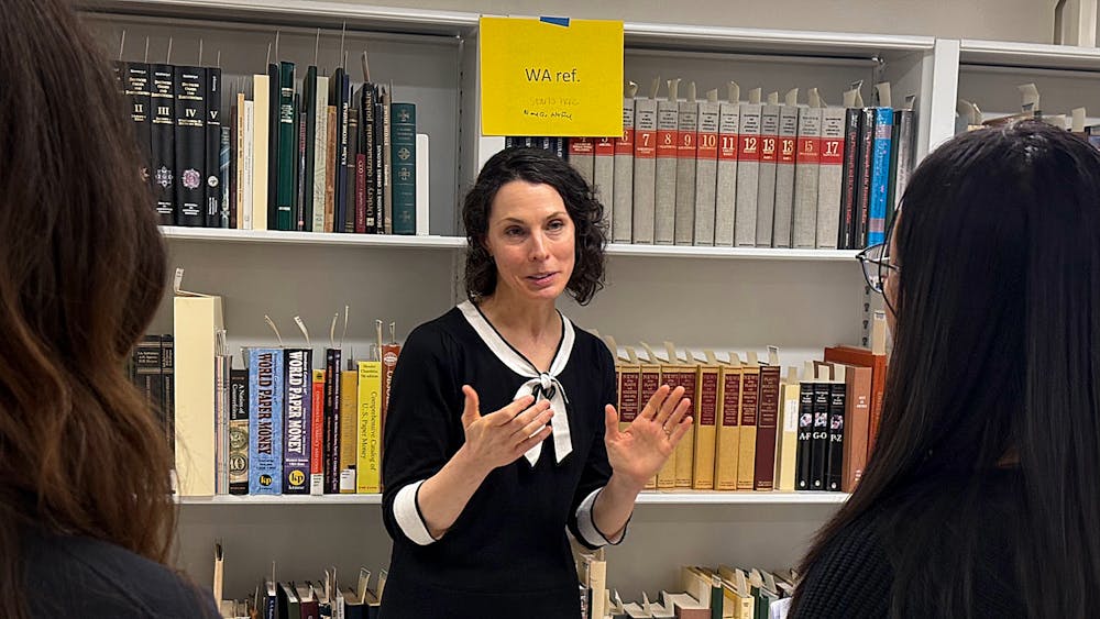 A woman with short dark hair wearing a black shirt with white trim stands before shelves of books, speaking with two women who stand facing her.