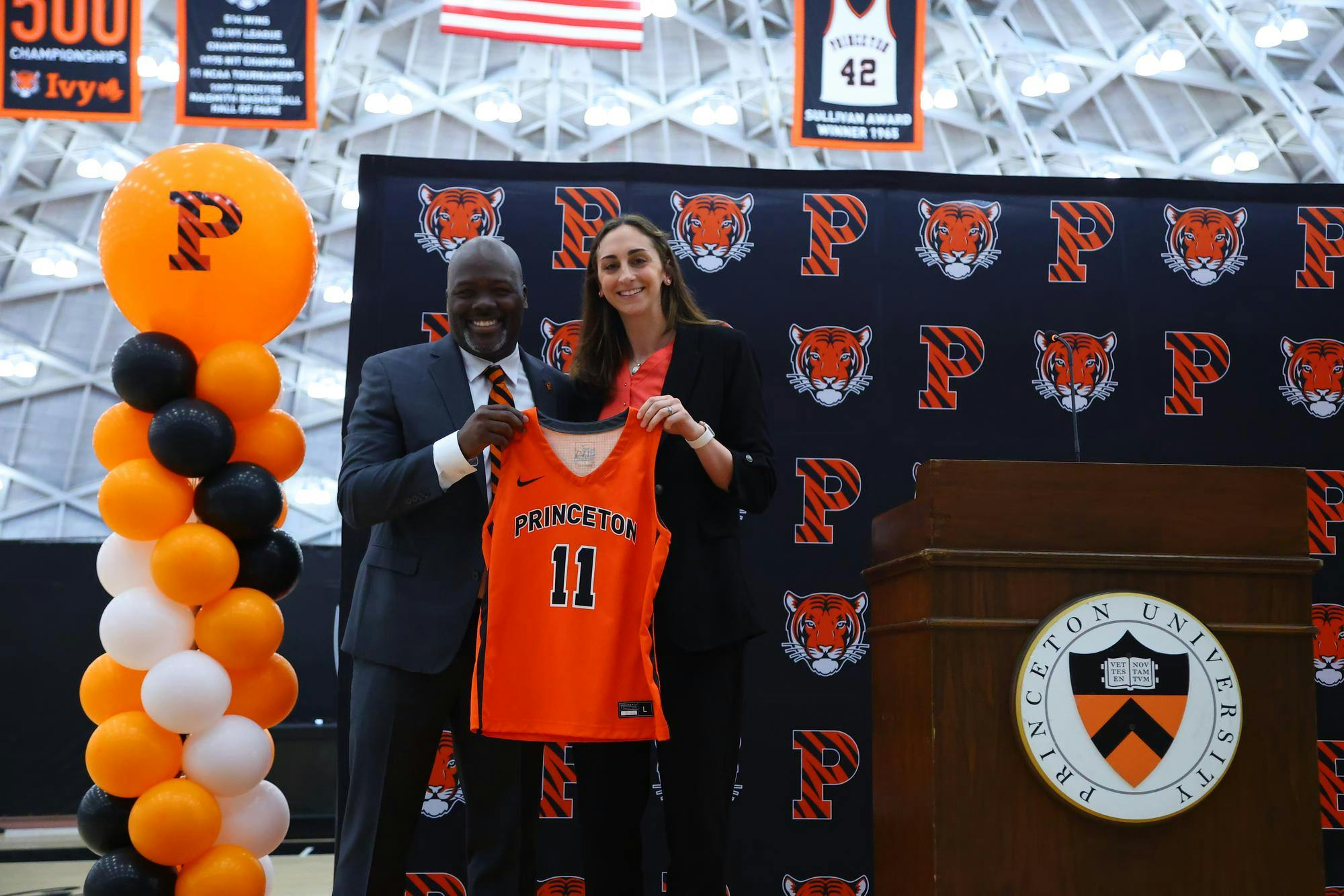 The head coach stands next to the athletic director at an introductory event, holding up an orange Princeton jersey with the number 11 in front.