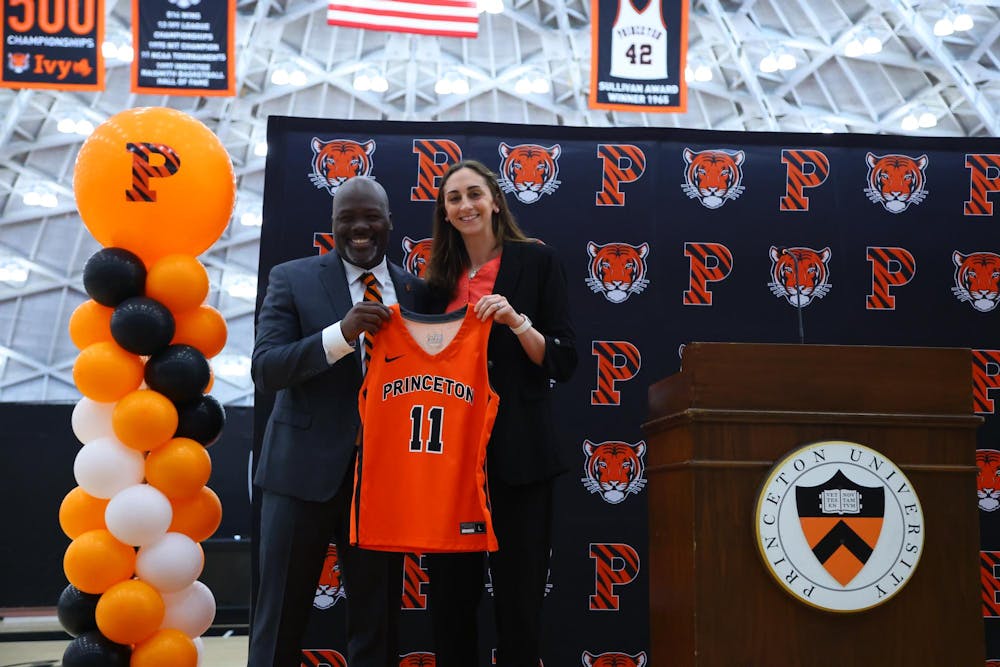 The head coach stands next to the athletic director at an introductory event, holding up an orange Princeton jersey with the number 11 in front.