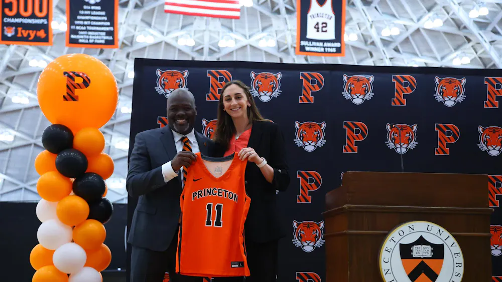 The head coach stands next to the athletic director at an introductory event, holding up an orange Princeton jersey with the number 11 in front.