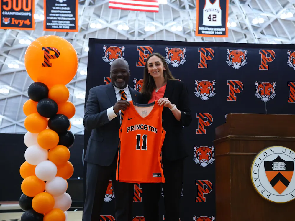 The head coach stands next to the athletic director at an introductory event, holding up an orange Princeton jersey with the number 11 in front.