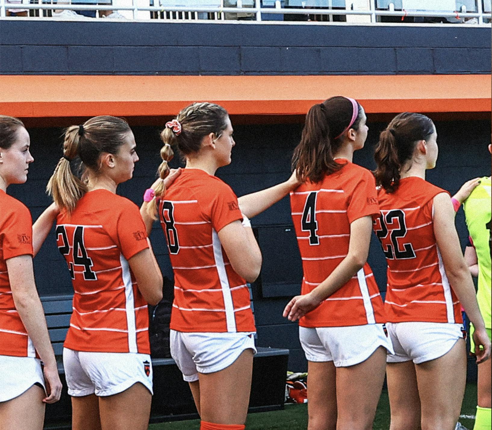 Women's soccer players standing in line with each girl's left arm on the left shoulder of the girl in front of them